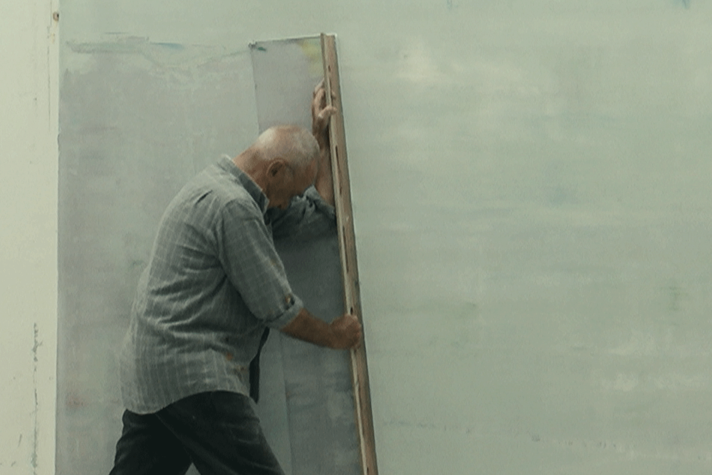 The artist Gerhard Richter pushes a large vertical squeegee across an oversized green-tinted off-white canvas to reveal gray off-white paint layers underneath
